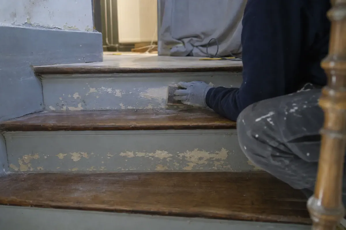 Compagnon ponçant la contremarche écaillée d’un escalier en bois d’immeuble ancien, marches cirées