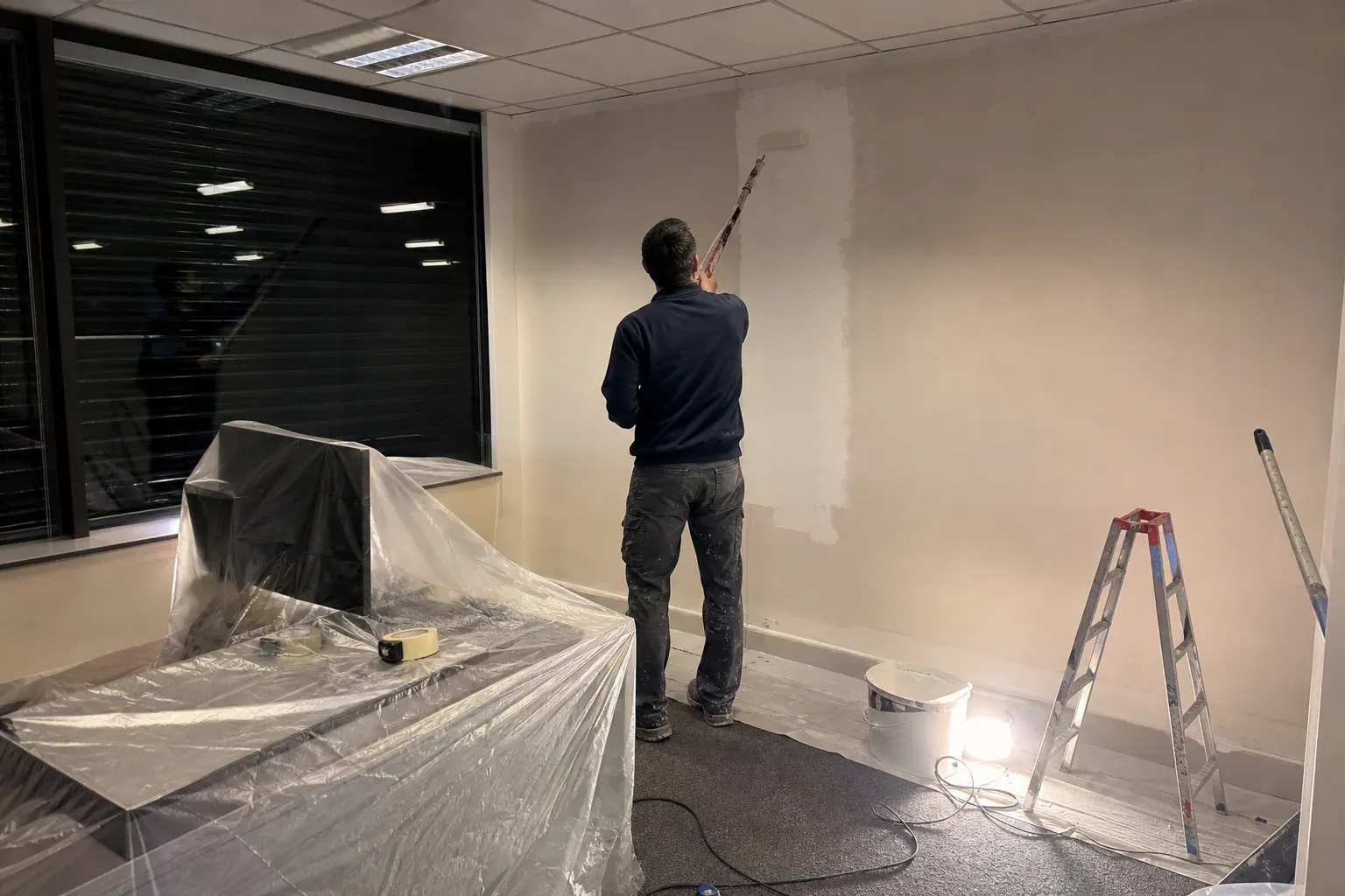 Peintre repeignant la cloison d’un bureau de nuit, postes de travail couverts de bâches