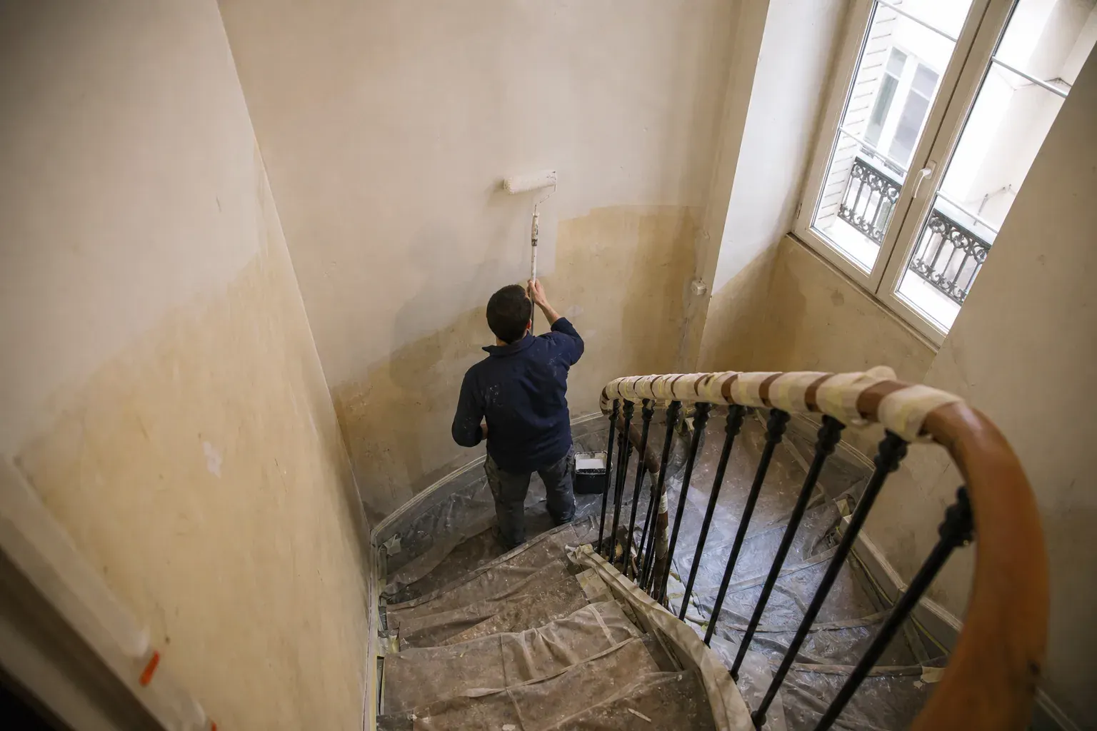 Peintre de dos repeignant une cage d’escalier d’immeuble étage par étage, marches bâchées