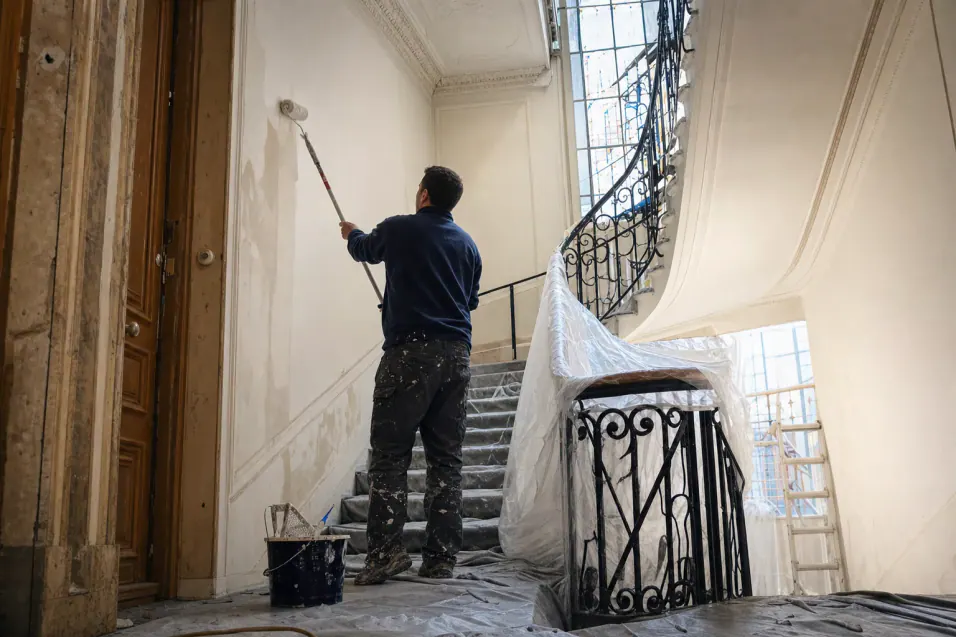 Peintre en bâtiment de dos repeignant le palier d’une cage d’escalier haussmannienne, rampe en fer forgé protégée