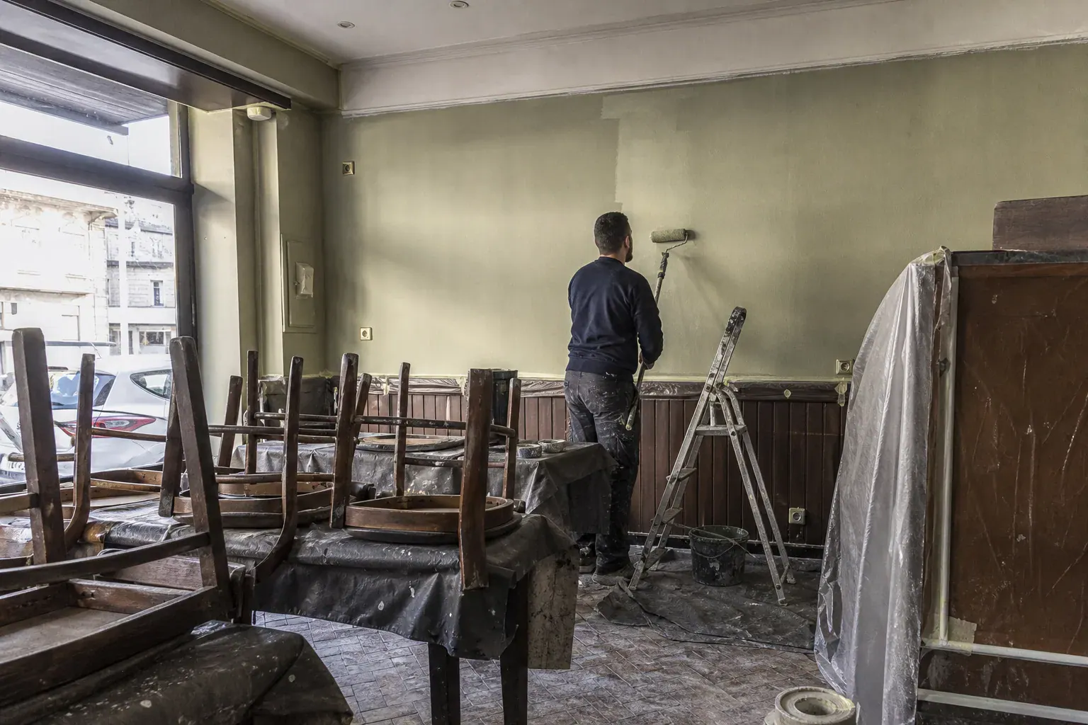 Peintre repeignant en vert sauge la salle d’un restaurant du Val-de-Marne, chaises retournées sur les tables bâchées