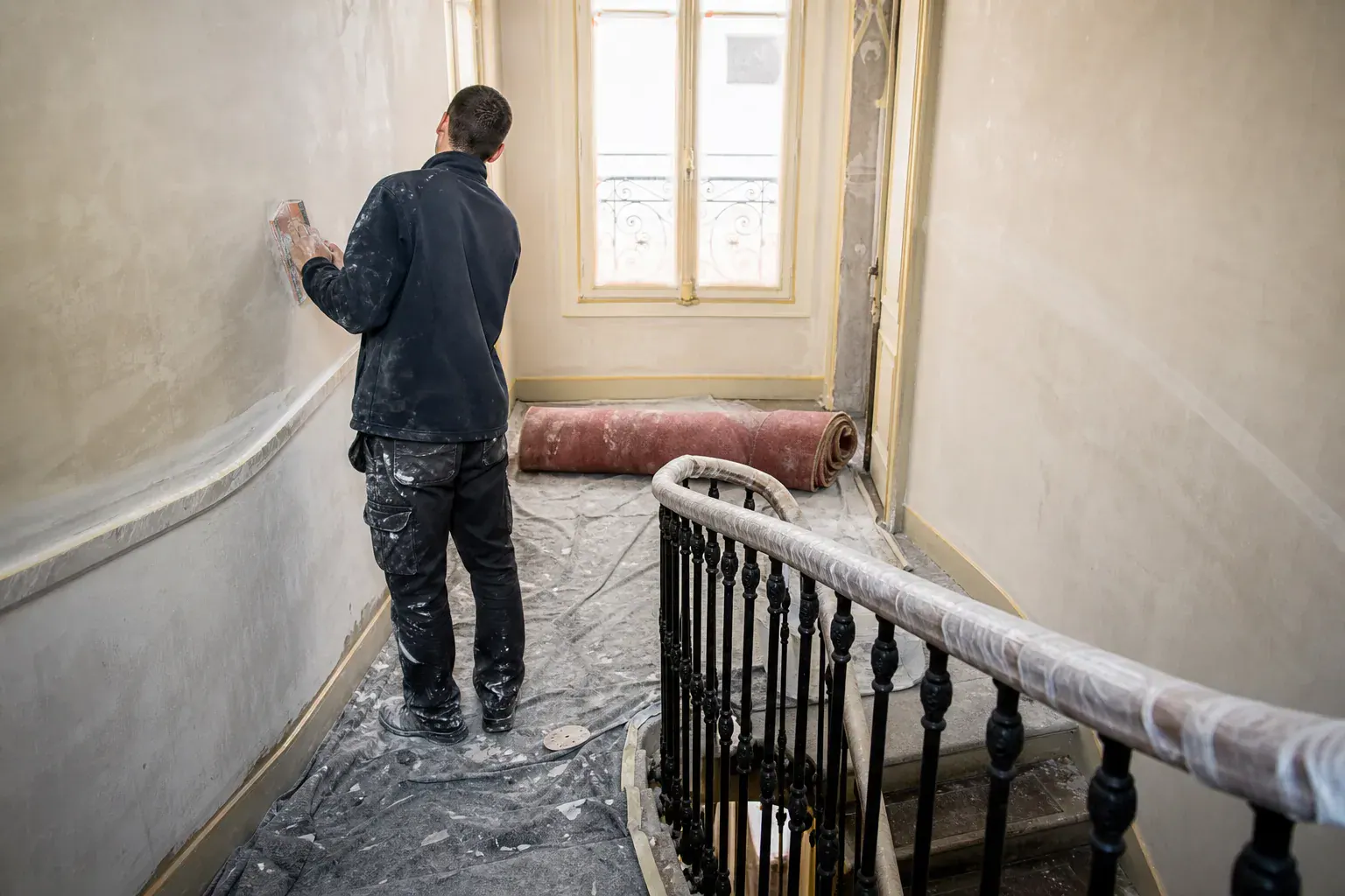 Compagnon ponçant le plâtre d’une cage d’escalier haussmannienne à Paris, tapis roulé et rampe en fer forgé gainée