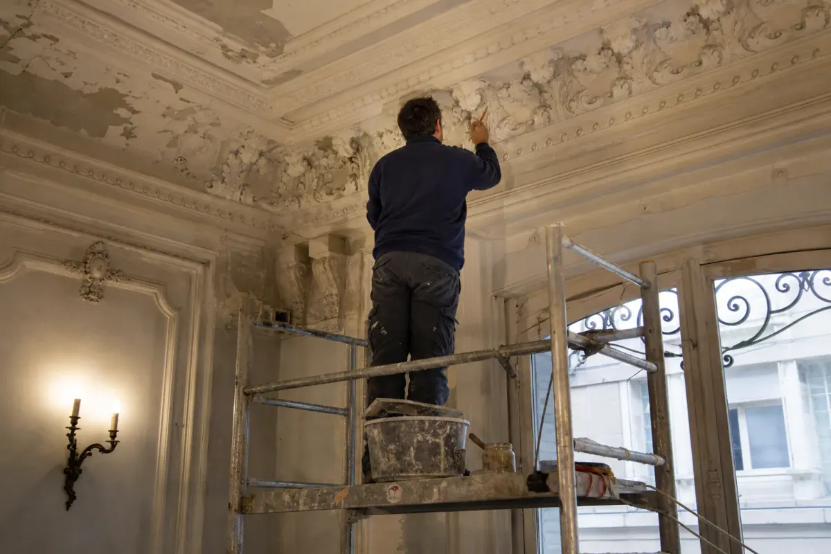 Peintre reprenant au pinceau les moulures en staff d’un hall haussmannien sans les empâter